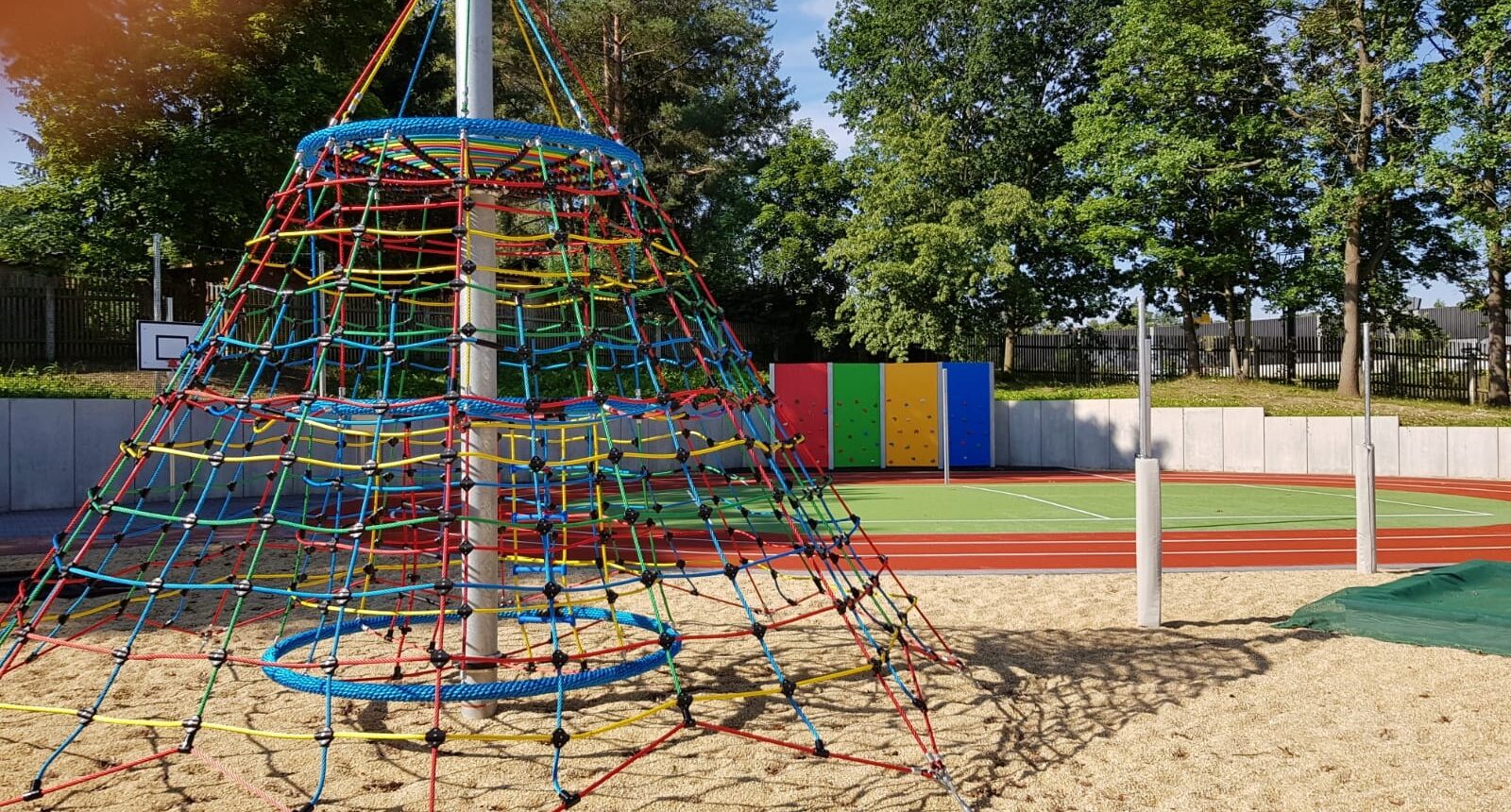 Der Spielplatz im Hof. Im Vordergrund steht eine Kletterspinne in Kegelform, rechts im Bild sind zwei Pfosten, an denen eine Schnur oder ein Volleyballnetz befestigt werden kann. Im Hintergrund sieht man eine Mini-Laufbahn, mit einem Mini-Fußballfeld darin. Dahinter befindet sich eine Kletterwand und links neben der Kletterwand steht ein Basketballkorb.