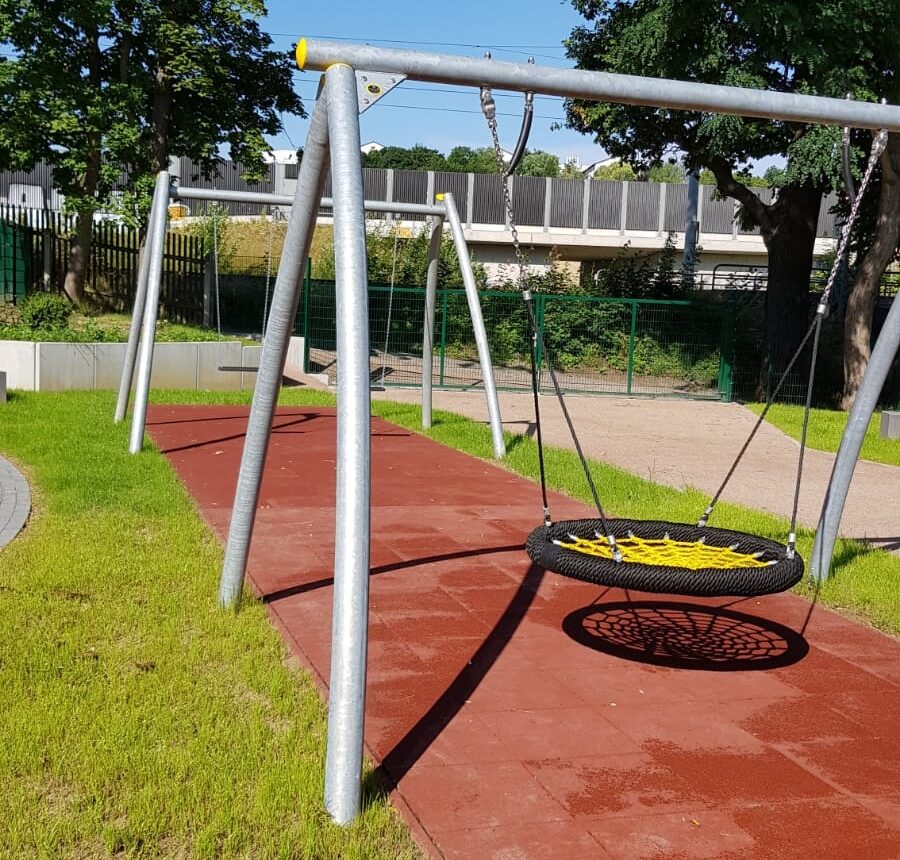 Auf dem Bild ist im Vordergrund eine Vogelnestschaukel zu sehen. Der Außenrand der Schaukel ist schwarz und die Innenseile sind gelb. Unter der Schaukel sind gummierte Bodenplatten verlegt, welche die Schüler bei einem Sturz schützen sollen. Die Platten sind so weit verlegt, dass auch die beiden normalen Schaukeln im Hintergrund über den Platten stehen.