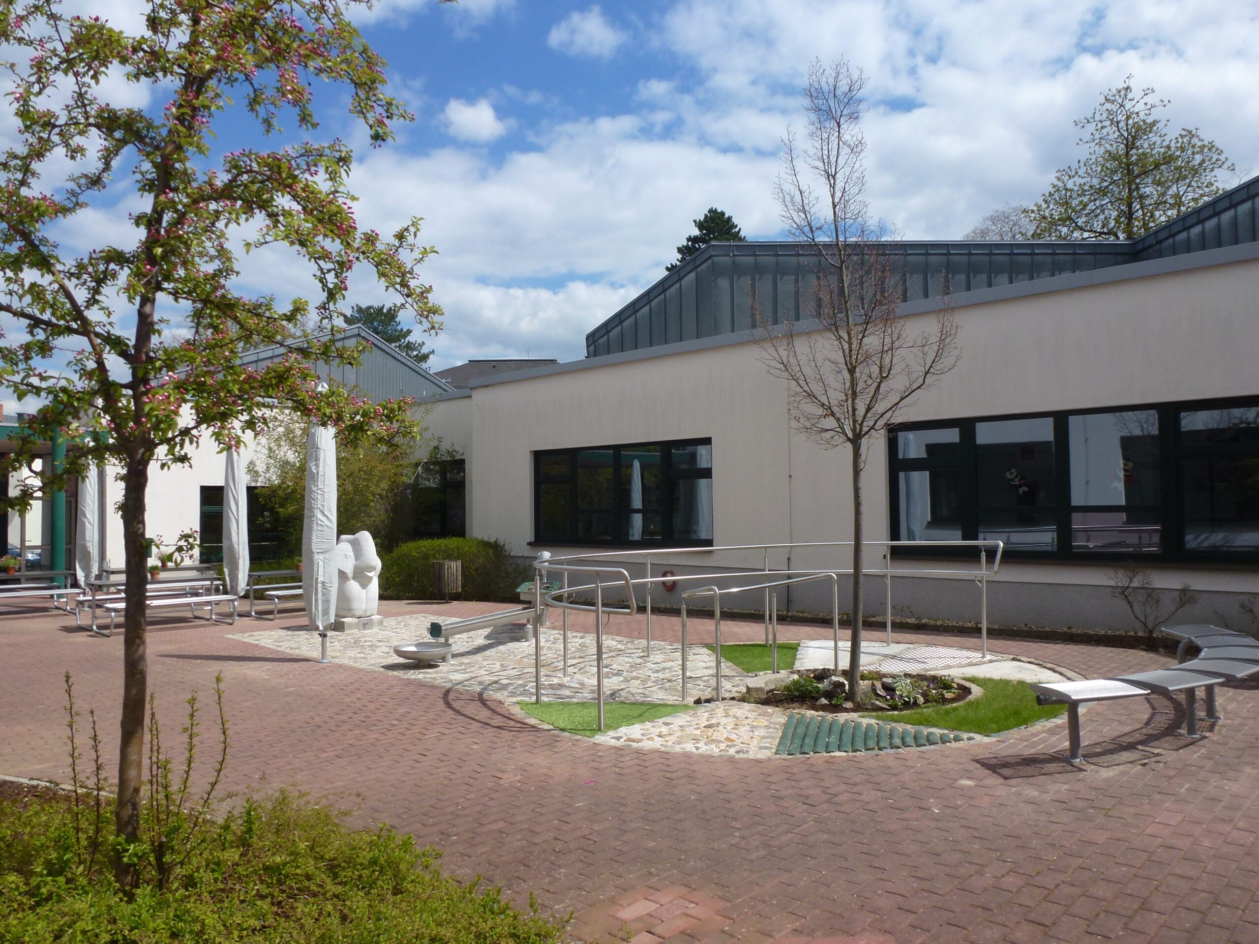 Der Hof der Sonnenbergschule. In der Mitte des Hofes ist ein Wasserspielplatz und eine Barfußstrecke. Daneben befindet sich die Statue des kleinen Elefants. Hinter dem Elefant ist das grüne Klassenzimmer und zwischen den Bänken des grünen Klassenzimmers stehen Sonnenschirme.