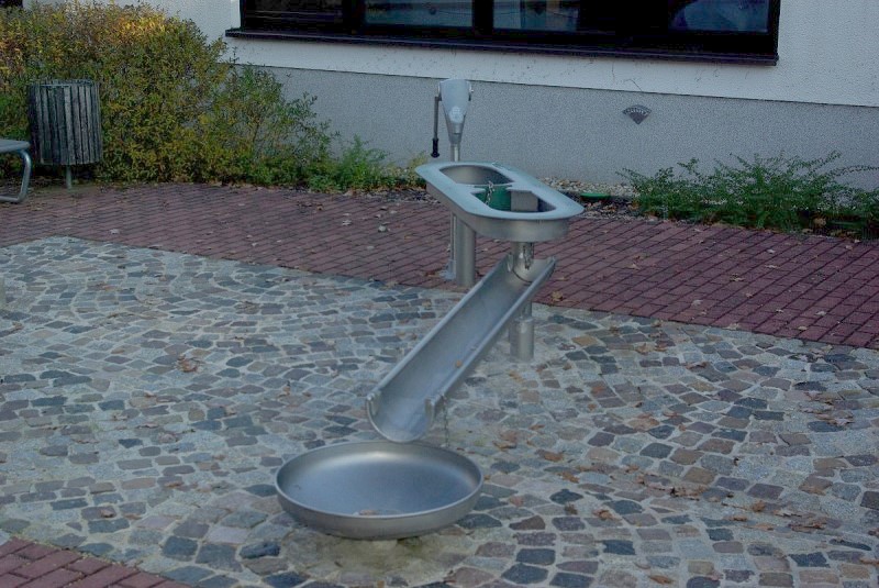 Das Wasserspiel auf dem Schulhof. Über eine Pumpe kann Wasser gefördert werden, dieses läuft dann in ein kleines Becken. Von diesem Becken aus kann das Wasser mit einem kleinen Eimer oder der eingebauten Sperre in das Becken daneben geleitet werden. Von diesem Becken fließt das Wasser in eine Rinne und dann in eine Schale, wo es durch einen Abfluss wieder im Boden versinkt.