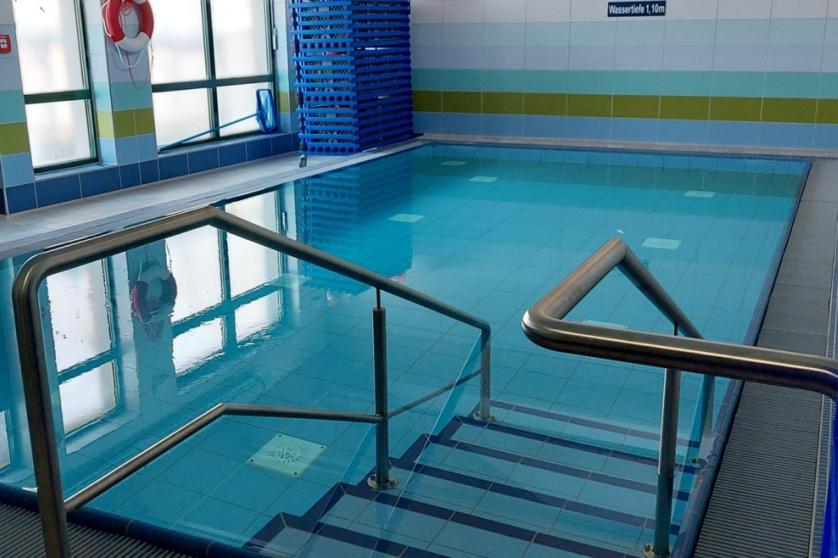 Das Schwimmbad der Schule. Das Foto wurde von der Einstiegstreppe aus aufgenommen. Links im Bild sieht man bodentiefe, milchige Fenster, einen Rettungsring an der Säule zwischen den Fenstern und einen Kescher. Rechts im Bild sieht man die geflieste Schwimmbadwand.