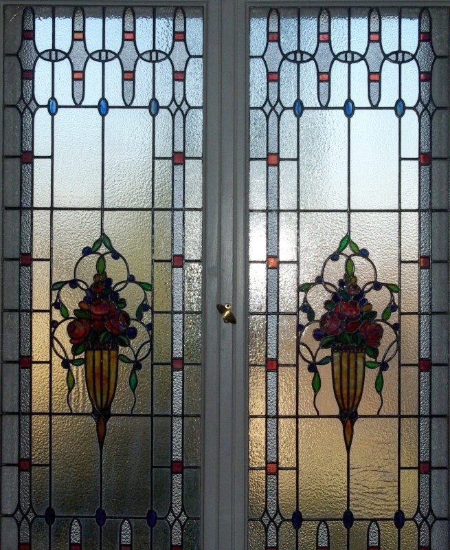 Ein Fenster im Jugendstil mit zwei Fensterflügeln. Die beiden Fensterflügel sind gespiegelt. In der Mitte der Fenster befindet sich die Darstellung einer Blumenvase mit roten Blumen