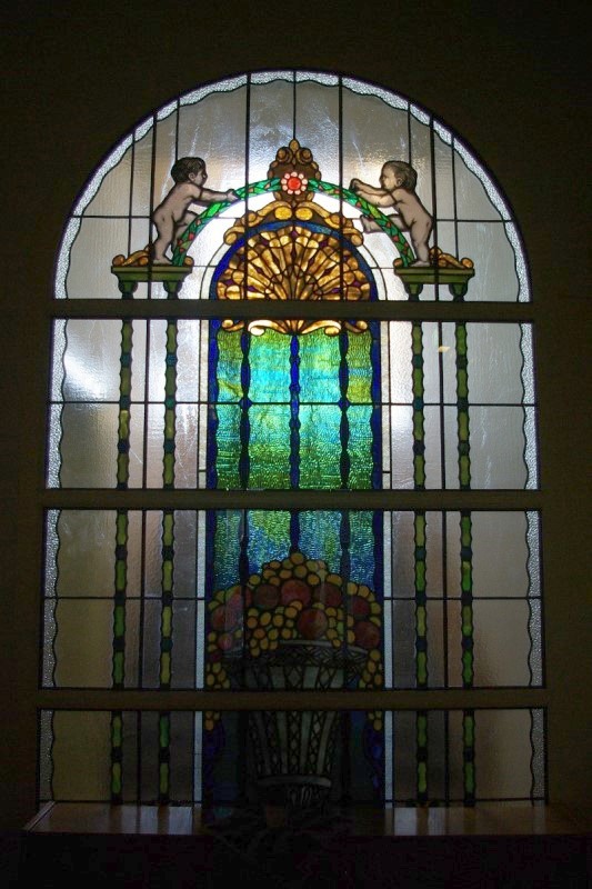 Ein Buntglasfenster im Jugendstil. Unten ist eine hohe Vase mit bunten Früchten zu sehen, Rechts und links von der Vase gehen zwei Säulen nach oben. Auf den Säulen steht jeweils ein Knabe, welcher an dem Bogen über der Vase hinaufklettern will.
