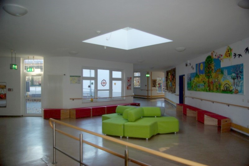Der Neubau. In der Mitte des Neubaus ist in der Decke ein Fenster eingelassen, wodurch der ganze Raum hell wirkt. Unter dem Deckenfenster steht ein grünes Sitzmöbel. An den Wänden stehen Bänke und es führt ein Handlauf entlang. Über dem Handlauf ist ein großes Wandgemälde mit den vier Jahreszeiten.