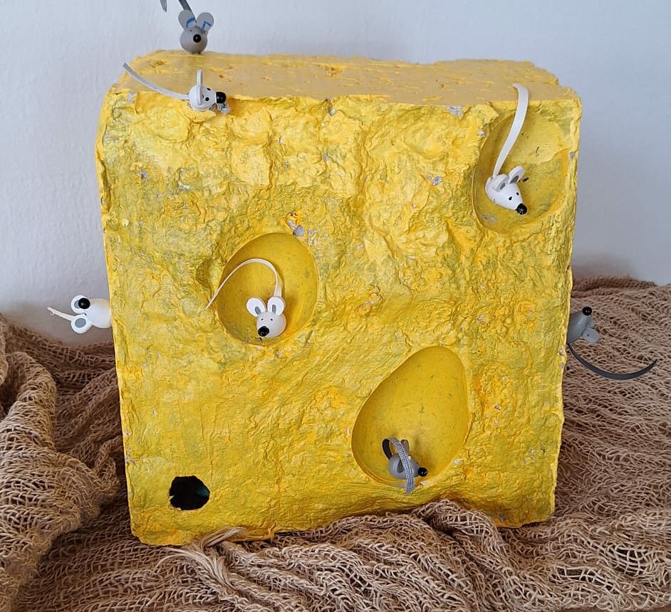 Ein gelb angemalter Betonblock, welcher kleine Kuhlen ausgeschnitten hat. Der Betonblock sieht somit aus wie ein Stück Käse. In den den Kuhlen befinden sich kleine Mäusefiguren.