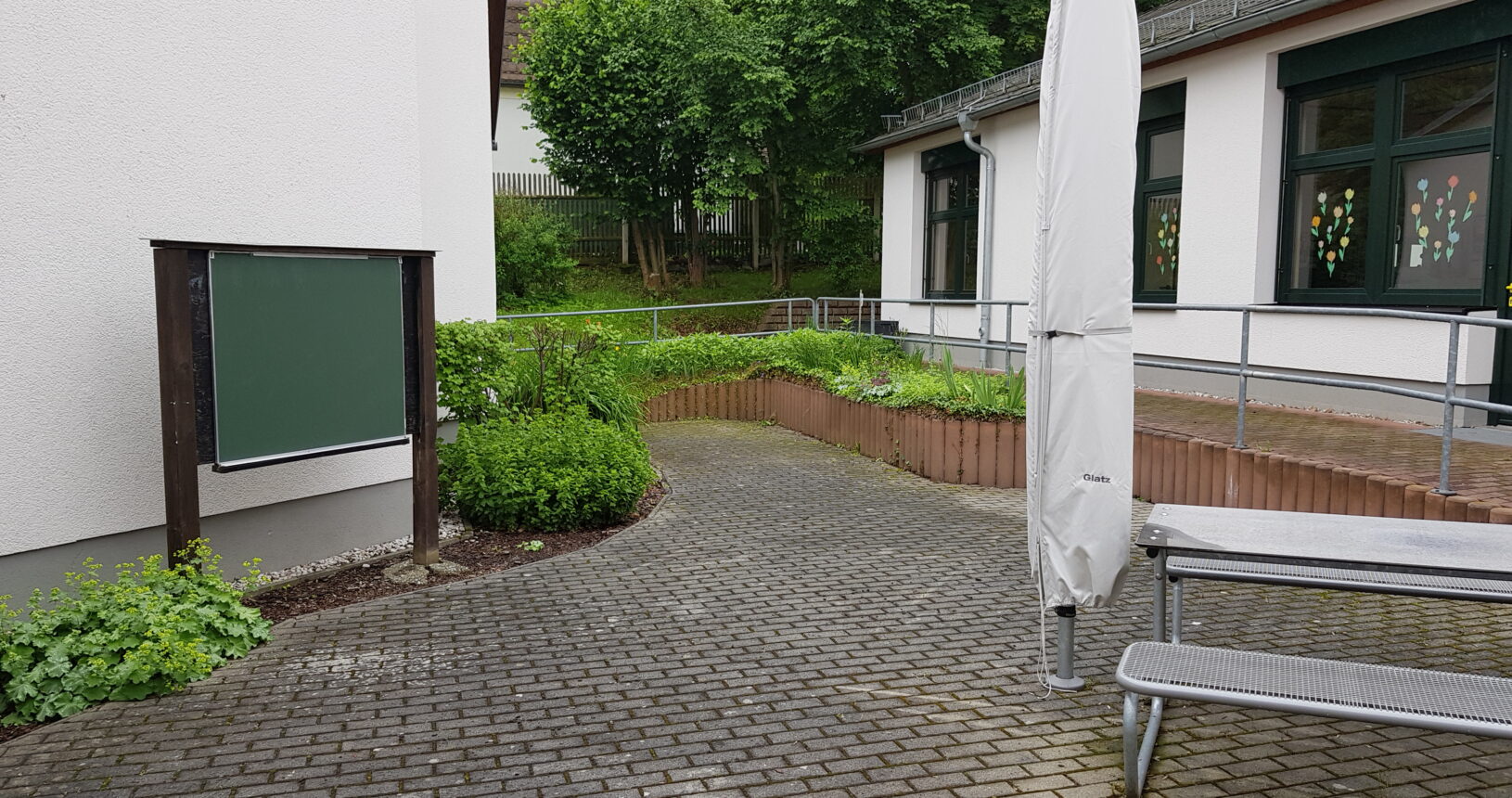 Ein Bild des grünen Klassenzimmers. Auf der linken Seite neben der Wand des Schulgebäudes steht eine Tafel, auf der rechten Seite sind die Sitzbänke und Tische für die Schüler.