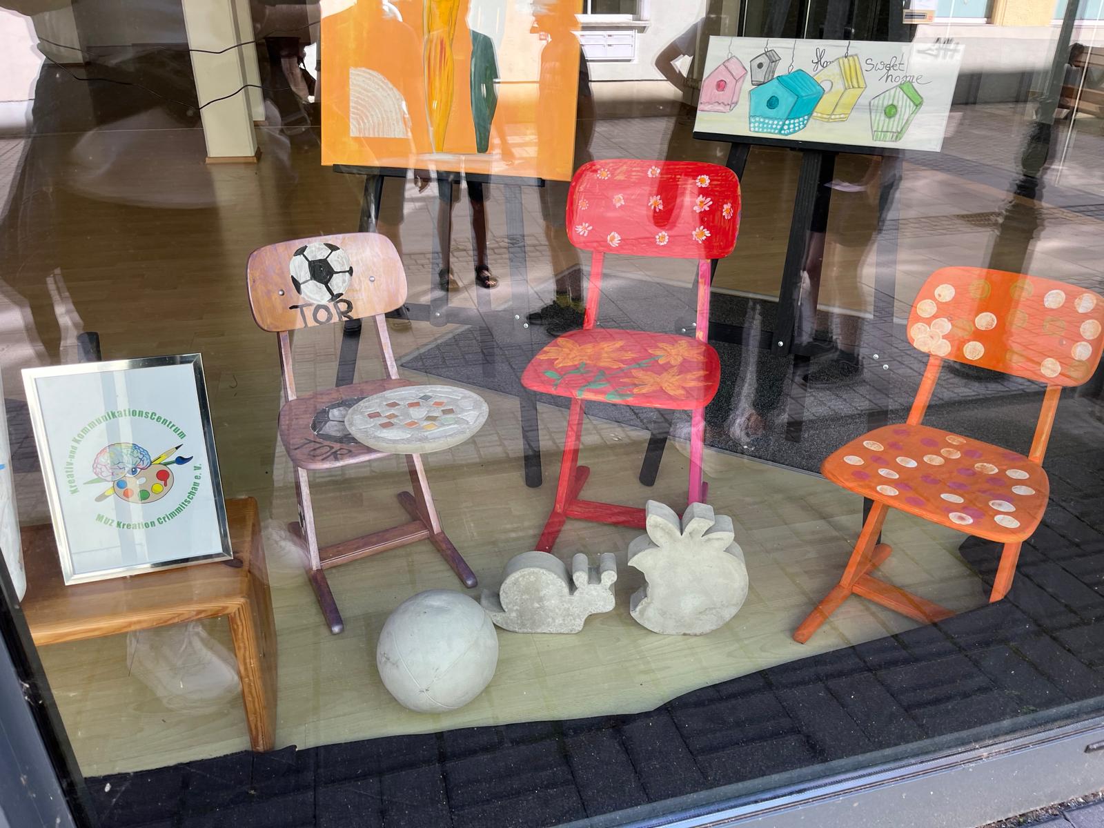 Die Gestaltung eines Schaufensters im rahmen des Projektes "Kunst im öffentlichen Raum." Zu sehen sind drei bemalte Stühle, zwei Gemälde und drei Betonfiguren (eine Schnecke, eine Kugel und ein Apfel).