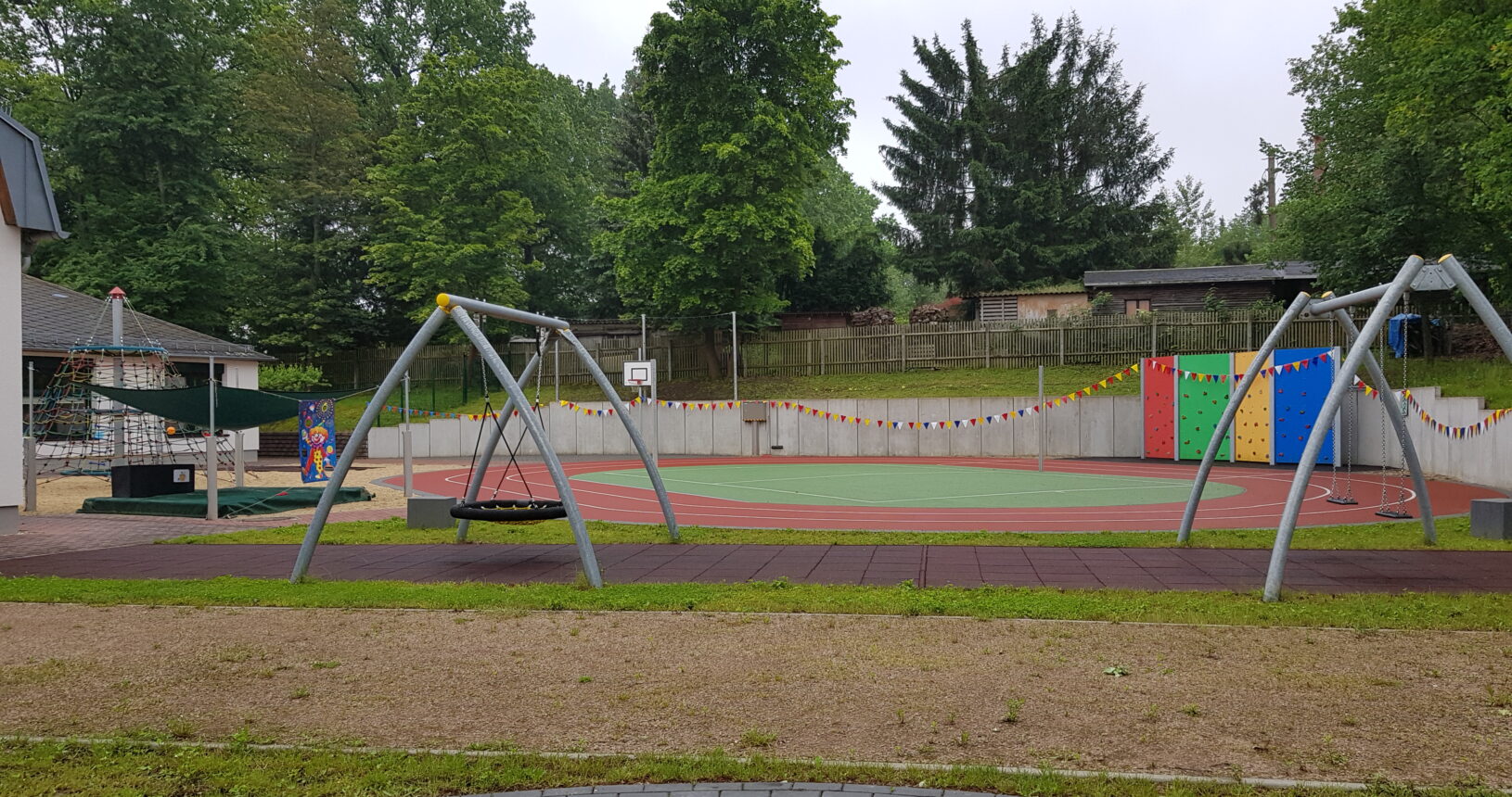 Ein Bild des Spielplatzes. Auf der linken Seite sieht man noch die Kletterspinne. Im Fokus des Bildes hingegen befinden sich die Schaukeln. Die Schaukel auf der linken Seite ist eine Vogelnestschaukel, die beiden Schaukeln auf der rechten Seite sind normale Schaukeln. Im Hintergrund sieht man wider den Sportplatz.