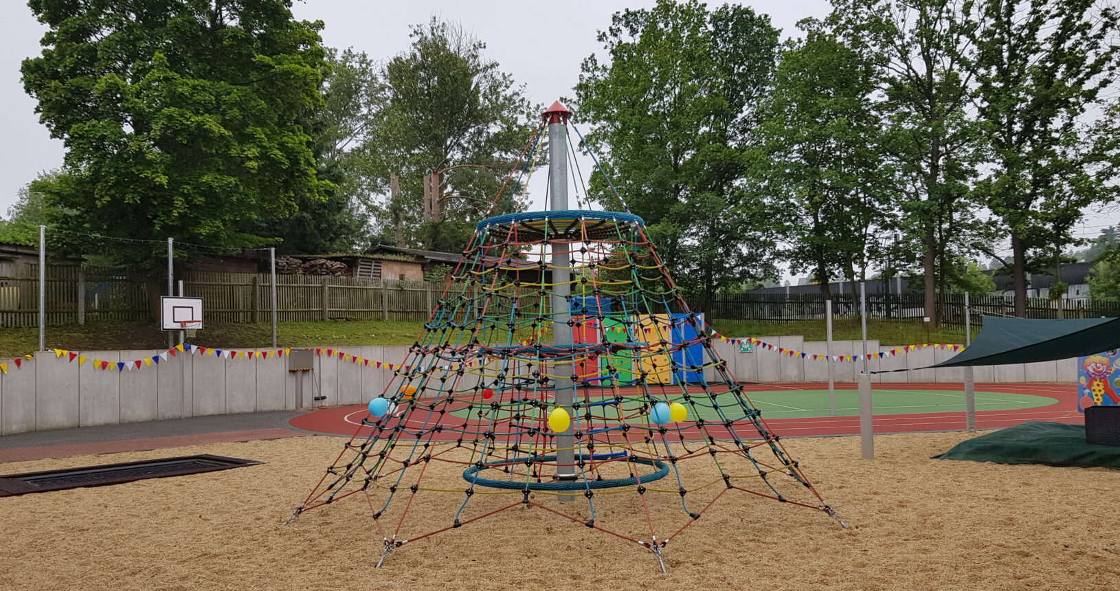 Ein Bild des Spielplatzes. Im Fokus steht hierbei die große Kletterspinne, dahinter kann man den Sportplatz erkennen.