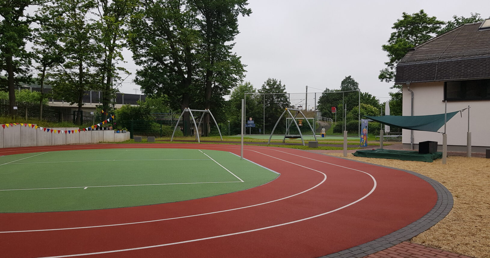 Der Sportplatz der Schule. Dieser besteht aus einer Laufbahn und einem grünen Kunstrasenfeld in der Mitte der Bahn. Auf der rechten Seite kann man einen, mit einem Sonnensegel überdachten Sandkasten des Spielplatzes sehen. Im Hintergrund sieht man die Schaukeln.